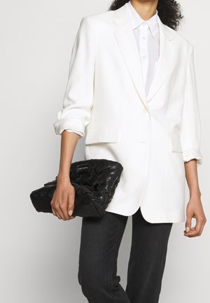 White tailored blazer over a white shirt, paired with a black quilted clutch bag featuring a logo accent. Dark jeans complete the outfit.