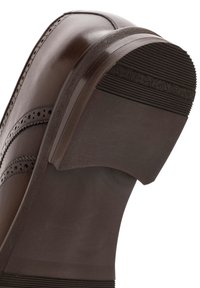 Chaussure habillée en cuir marron avec perforations décoratives, montrant une semelle lisse et un talon texturé, inclinée pour afficher la partie inférieure.