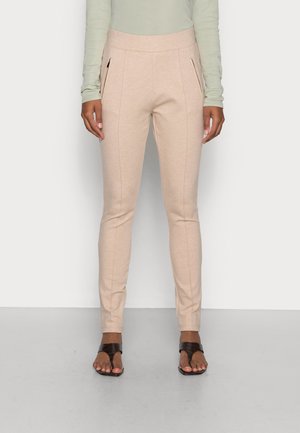Pantalon beige slim fit avec poches latérales zippées, porté avec un haut manches longues vert clair et des sandales noires à bout ouvert.