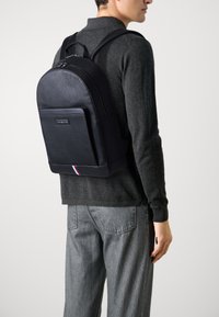 Sac à dos en cuir noir avec surface texturée, haut arrondi, poche avant et accent rayé en bas. Le modèle porte un pull gris foncé et un jean.