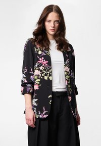 Young woman with wavy brown hair wearing black floral blazer over white top and black wide-leg pants against white background.