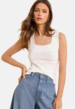 Hosszú barna hajú nő, fehér ujjatlan kötött felsőt és világoskék magasderekú farmert visel, kezét a csípőjén tartva pózol.