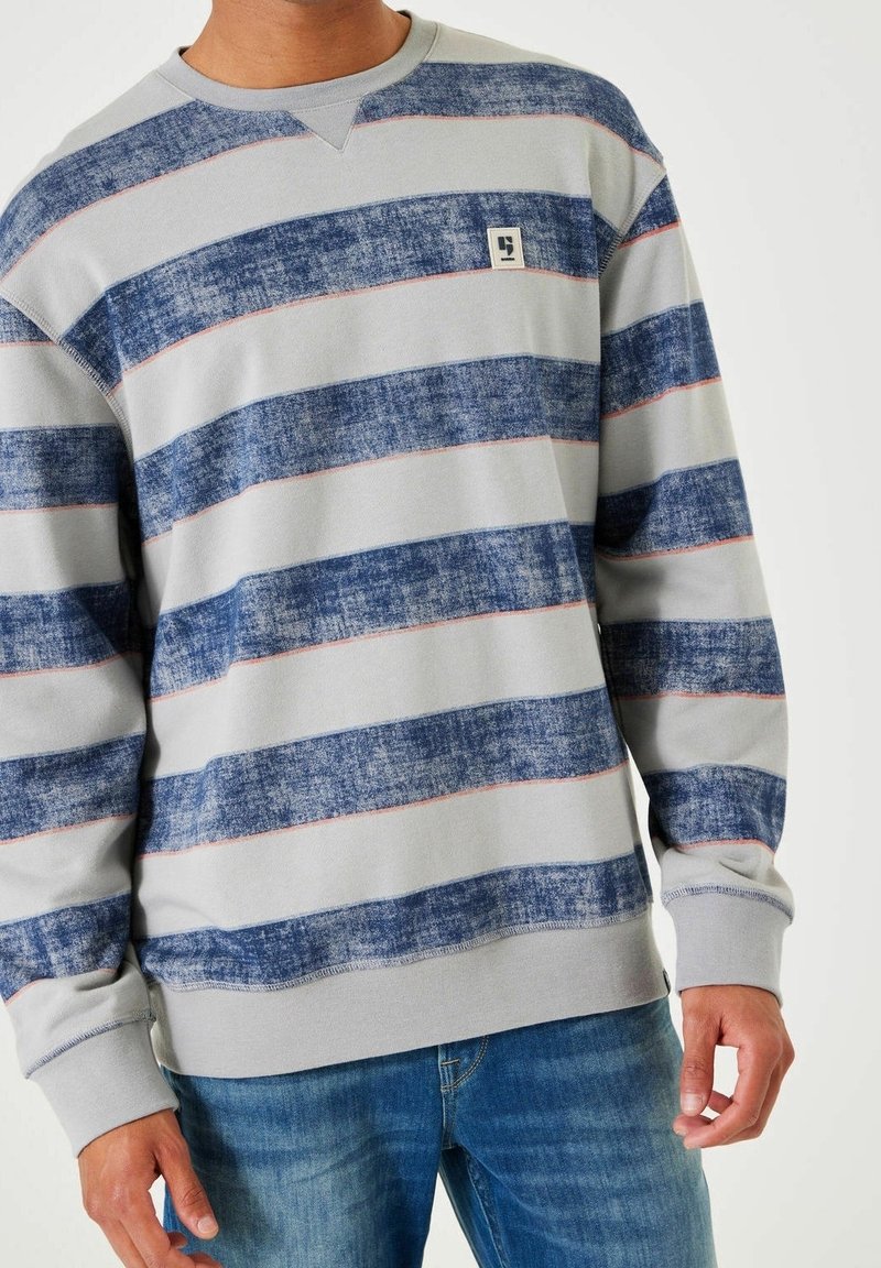 Homme portant un sweat-shirt gris à manches longues avec de larges bandes horizontales bleues texturées et un petit logo sur la poitrine.
