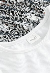 Vit ribbad t-shirt med rundade halsringning. Designen har svarta och silverfärgade paljetter på den övre delen, vilket skapar en glittrande effekt.
