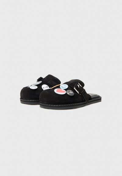 Zapatos de ante negros tipo mocasín con puntera redondeada, que presentan múltiples botones decorativos de diferentes colores y diseños, y una correa con hebilla.