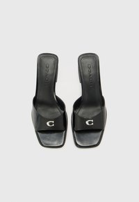 Sandalias de cuero negro sin talón con una tira ancha, que presenta un logo plateado en forma de "C". Diseño de puntera cuadrada y parte trasera abierta, textura suave.