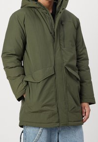 Oliivinvihreä nylonista valmistettu parka-takki, jossa on huppu, vetoketjullinen etutasku ja etutaskut läpällä, jotka ovat teksturoidulla pinnalla.