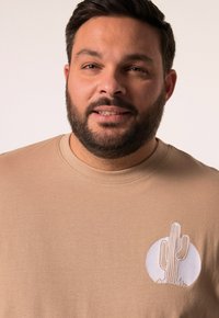 Béžové bavlněné tričko s malým bílým vyšitým designem kaktusu na levé části hrudi. Kulatý výstřih a krátké rukávy.