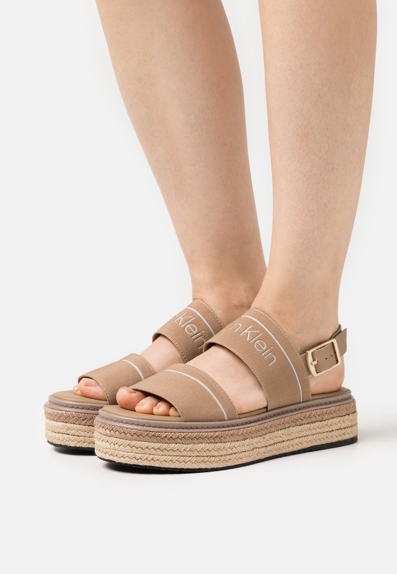 Pieds portant des sandales compensées beiges avec des semelles tissées épaisses et des brides en tissu de marque sur les orteils et les chevilles avec une fermeture à boucle.