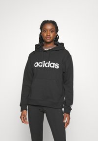 Črna kapuca s puloverjem z velikim belim logotipom "adidas", sprednjim žepom in rebrastimi manšeti. Uporabljena z črnimi leginami. Gladka tekstura tkanine.