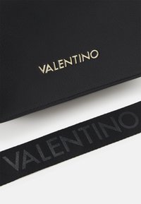 Czarna torebka z teksturowanej skóry z złotym logo "VALENTINO". Posiada szeroki czarny materiałowy pasek z tłoczonym napisem. Gładkie wykończenie, minimalistyczny design.