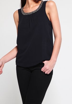 Frau trägt ein ärmelloses schwarzes Oberteil mit einem perlenbesetzten Ausschnitt, kombiniert mit schwarzen, eng anliegenden Hosen, steht vor einem schlichten Hintergrund.