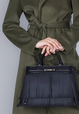 Mustasta nahkakäsilaukusta, jossa on koristetupsut, pyöreät kahvat ja kultasävyinen logon yksityiskohta. Laukku pidetään oliivinvihreän takin vasten.