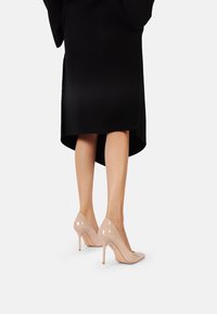 Tacchi alti in pelle verniciata beige con punta affusolata e tacco a stiletto sottile, abbinati a un cappotto nero fino al ginocchio.