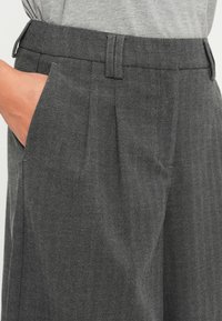 Pantalons en chevrons gris présentant un devant plissé, des poches latérales et une texture lisse. La taille est plate avec des passants de ceinture.