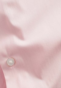 Helles rosa Gewebe mit glatter Textur und einem einzelnen weißen Knopfdetail. Der Stoff ist leicht zerknittert und zeigt Drapierungen.
