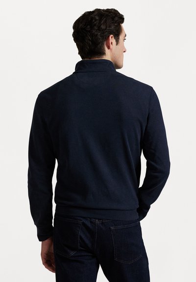 Mørkeblå genser laget av myk stoff, med høy krage og lange ermer, paret med mørke denimbukser. Glatt tekstur, ingen mønstre.