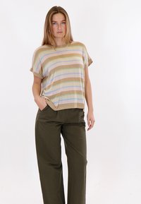 Woman standing with one hand in pocket, wearing a short-sleeve striped beige, green, blue top and olive green wide-leg pants on white background.