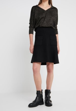 Kobieta w błyszczącej ciemnej bluzce, czarnej spódnicy do kolan z przednimi kieszeniami oraz czarnych botkach z klamrami na białym tle.