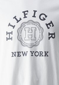 Tela blanca con texto negro "HILFIGER" curvado sobre un emblema circular que contiene una gran "H" y "COLUMBUS AVENUE SINCE 1985," y "NEW YORK" debajo.