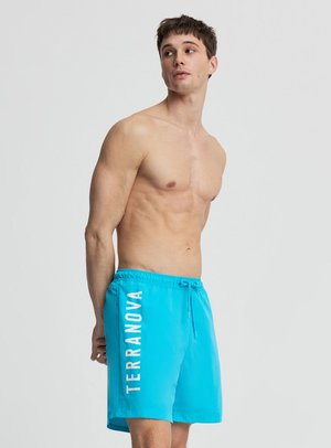 Uomo senza maglietta che indossa pantaloncini da bagno blu brillante con scritta bianca "TERRANOVA" sulla gamba sinistra, guarda verso sinistra su uno sfondo semplice.
