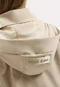 Jasno beżowa kurtka z kapturem, wykonana z gładkiego, matowego materiału. Posiada etykietę na szyi z wytłoczonym białym napisem "CROSS JEANS".