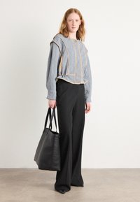 Gequilt bovenstuk in grijs met beige afwerking, gecombineerd met zwarte wijde broek. Het model houdt een zwarte leren tote bag vast. Soepele textuur en minimalistisch design.