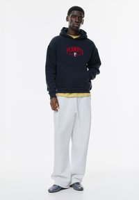 Hanorac navy cu logo-ul "ECHIPA DE GOLF PEANUTS" în roșu, suprapus peste un tricou galben. Asortat cu pantaloni de trening gri deschis și adidași gri.