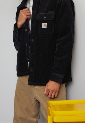 Personne portant une veste Carhartt en velours côtelé noir par-dessus une chemise blanche et un pantalon en velours côtelé marron clair debout près d’un contenant en plastique translucide jaune.