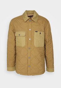 Hellbraune gesteppte Jacke mit klassischem Kragen, vorderer Knopfleiste und zwei Brusttaschen. Mit strukturiertem Stoff und kontrastierenden Ärmeln.