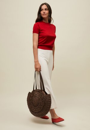 Woman in red short-sleeve top and white pants holding a large round woven brown bag, wearing red pointed flats, standing on neutral background.
