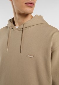 Felpa con cappuccio in misto cotone beige, con superficie strutturata e una patch del logo sulla parte anteriore. Inclusa chiusura con cordino.