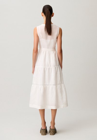 Donna con coda di cavallo lunga che indossa un vestito midi bianco senza maniche con gonna a balze e scarpe con tacco nere a motivo, vista di spalle su sfondo semplice.