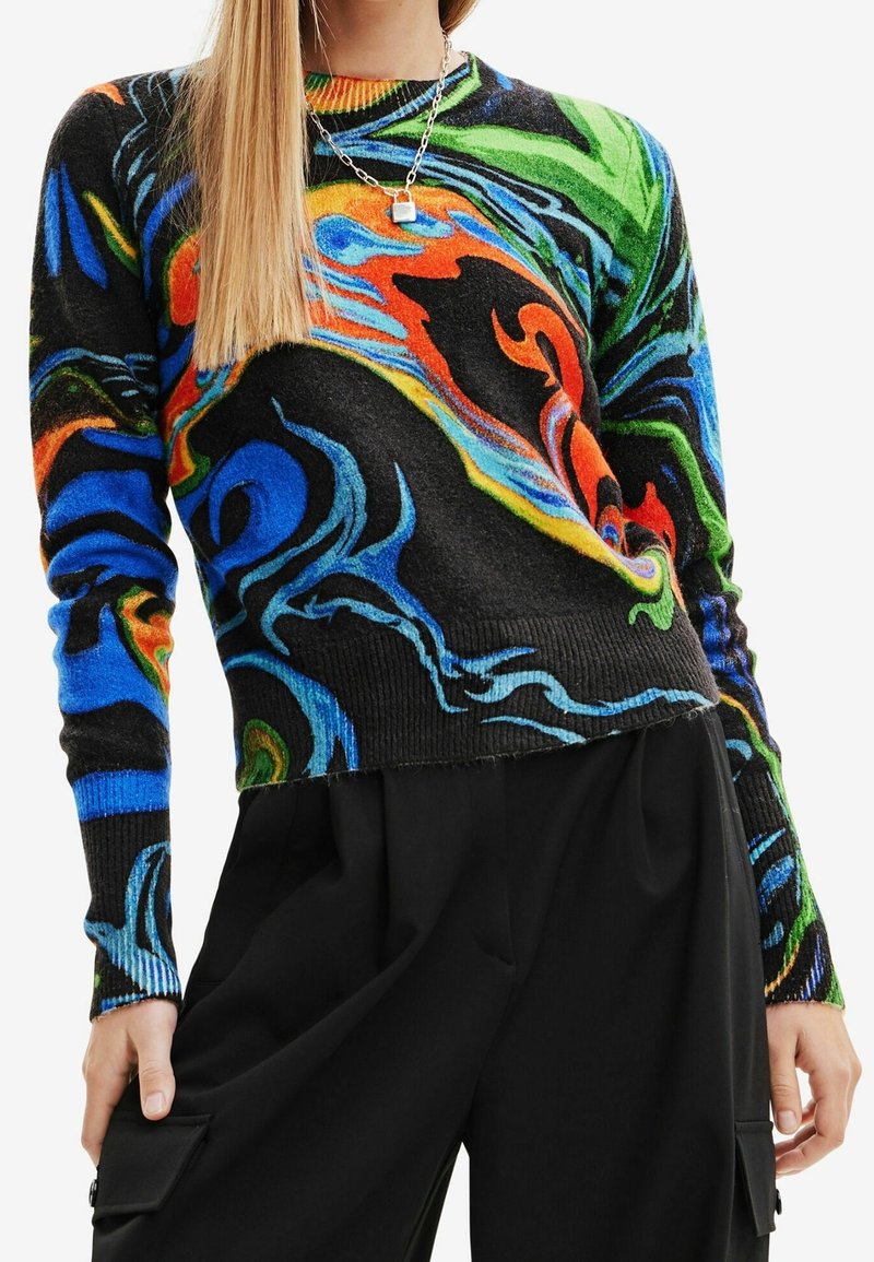 Femme portant un pull à motif abstrait multicolore, un pantalon noir et un collier chaîne argenté, tenant les rabats des poches des jambes de son pantalon.