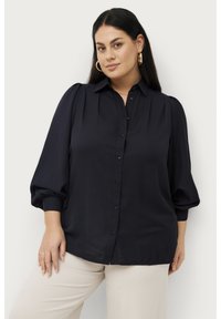 Marineblauw overhemd met opgestoken mouwen en een kraag, gemaakt van gladde stof. Subtiele plooien op de schouders, gecombineerd met lichtgekleurde pantalon.