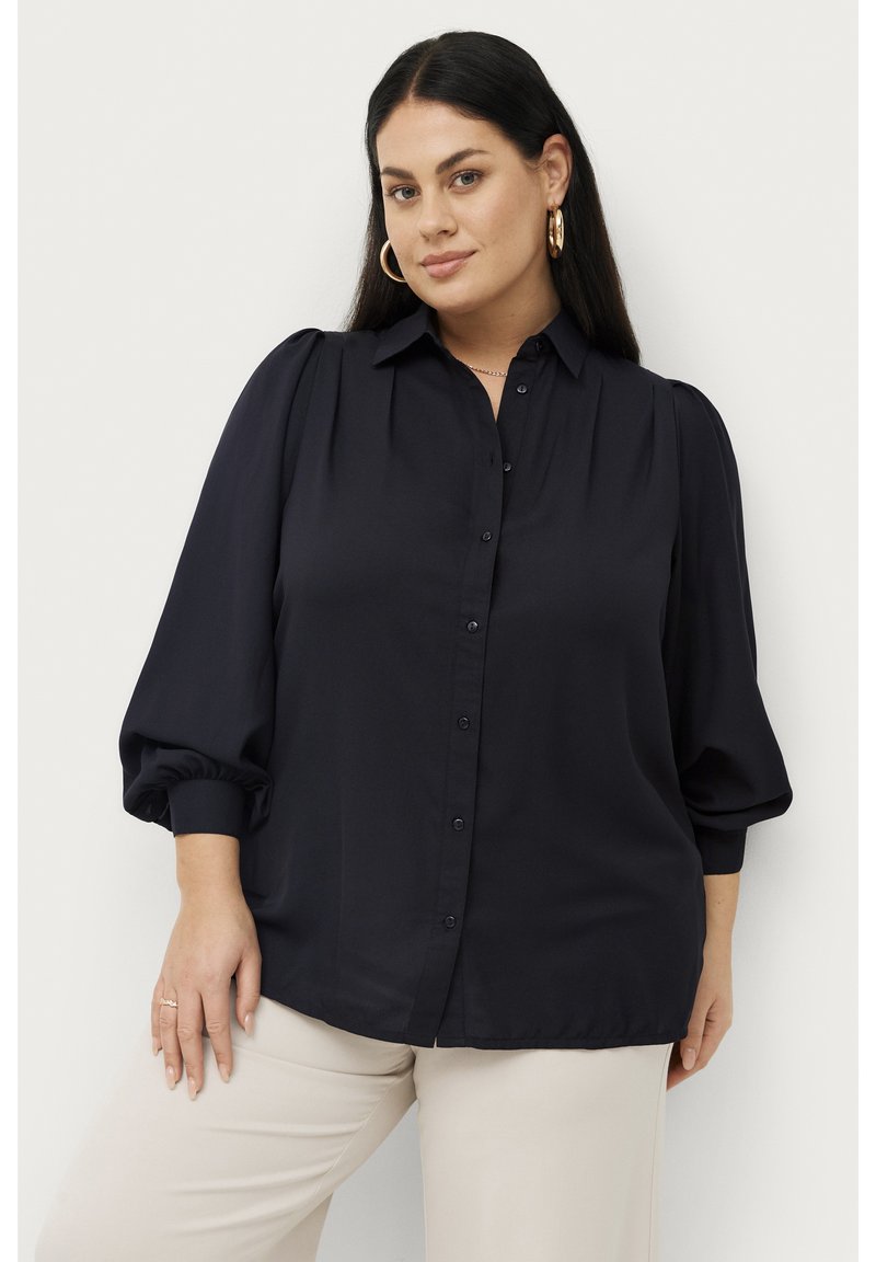 Marineblauw overhemd met opgestoken mouwen en een kraag, gemaakt van gladde stof. Subtiele plooien op de schouders, gecombineerd met lichtgekleurde pantalon.