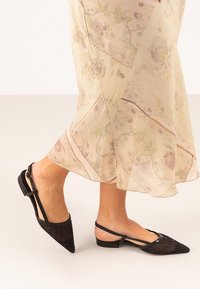 Zapatos negros de tipo slingback con punta afilada, con un diseño de malla tejida en la parte frontal, tacón plano y correa de tobillo ajustable.