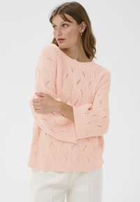 Femme portant un pull en tricot rose clair avec des motifs en vagues, un pantalon blanc et des boucles d'oreilles en or, posant les bras croisés et regardant à gauche.