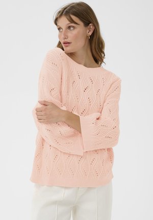 Frau in einem hellrosa Strickpullover mit Wellenmustern, weißen Hosen und goldenen Creolen, die mit verschränkten Armen posiert und nach links schaut.