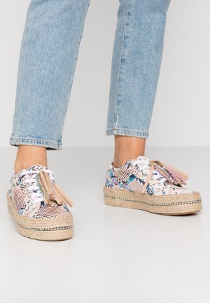 Pieds portant un jean bleu clair et des baskets compensées à motif serpent floral, avec des semelles tressées et des lacets à pompons.