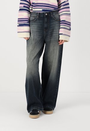 Wide-leg denim jeans in dark blue with a faded finish, featuring a high waist and five-pocket design, paired with black sneakers.