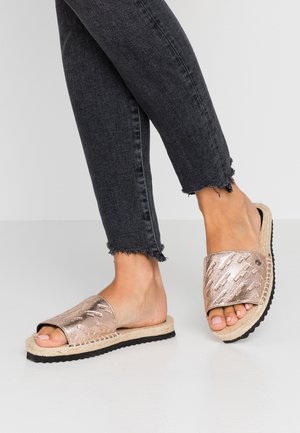 Stopy noszące metaliczne sandały typu slip-on z plecioną podeszwą i czarną gumową podeszwą zewnętrzną, zestawione z czarnymi, skróconymi dżinsami z postrzępionym dołem.