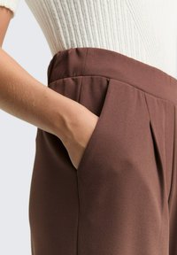 Brown trousers with a smooth texture, featuring side pockets and pleated detailing at the waist, paired with a ribbed white top.