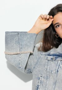 Ljusblå denimjacka med breda, fransade manschetter, med klassiska fickor och en blekt tvätt. Tyget har en strukturerad yta.