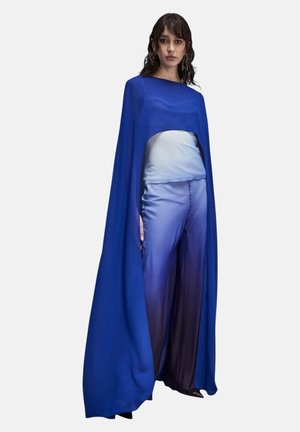 Donna che indossa un mantello fluente blu reale sopra una maglietta azzurro chiaro e pantaloni a gamba larga blu sfumati, in piedi su sfondo bianco.