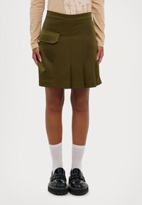 Olivengrønn plisséskjørt med en sidelomme, laget av et glatt materiale. Brukt med hvite sokker og blanke svarte loafers.