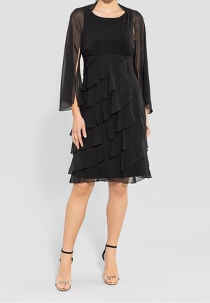 Nainen, jolla on päällään hihaton musta kerroksittainen mekko, jossa on läpikuultavat pitkät hihat, ja mustat nilkkahihna-sandaalit, seisoo yksinkertaisen vaalean taustan edessä.