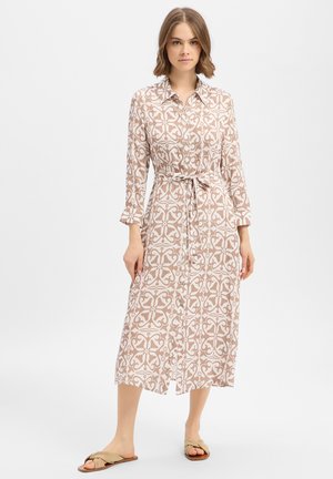 Frau trägt ein beige-weiß gemustertes, langärmliges Midikleid mit Gürtel und Knöpfen sowie beigen flachen Sandalen, stehend vor weißem Hintergrund.