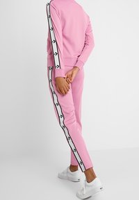 Pink tracksuit with white side stripes featuring black arrow patterns; model standing with back facing camera, wearing white sneakers.