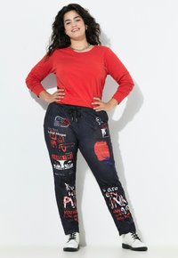 Rotes Langarmoberteil kombiniert mit schwarzen Jogginghosen, die verschiedene bunte Grafikdrucke aufweisen. Akzente sind eine verstellbare Taillenbindung und weiße Stiefel.